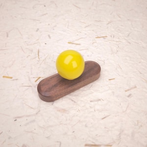 May include: A smooth, bright yellow spherical knob attached to a dark brown, elongated, oval-shaped wooden base. The object features a polished finish on the sphere and a natural wood grain texture on the base, creating a contrast between the two components.