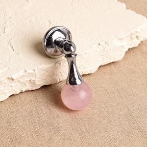 May include: A polished chrome cabinet knob with a teardrop-shaped pink quartz crystal. The knob has a round base and a curved neck, and the crystal is a soft, translucent pink. The knob is mounted on a light-colored, textured surface.