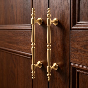 May include: Two gold-colored cabinet pulls with a classic design. Each pull has a long, slender handle with decorative detailing and is mounted on a dark brown wooden cabinet. The pulls are symmetrical and add a touch of elegance to the cabinetry.