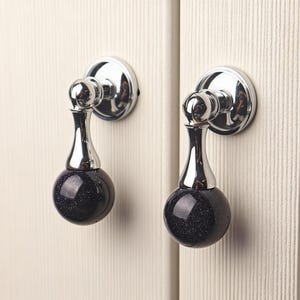 Könnte beinhalten: Zwei Schrankknöpfe mit polierter Chromoberfläche. Jeder Knopf hat eine dunkelblaue, gesprenkelte, kugelförmige Basis und einen dekorativen, länglichen Chromschaft. Die Knöpfe sind auf einer hellen, vertikal gerillten Oberfläche montiert.