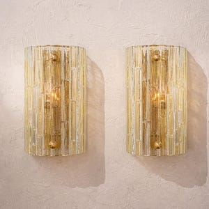 Op de afbeelding: Twee wandkandelaars met een textuur, cilindrisch ontwerp. De wandkandelaars zijn gemaakt van helder glas met verticale lijnen en een gouden tint. Elke wandkandelaar heeft een goudkleurige metalen armatuur en een warm licht dat van binnenuit komt.