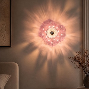 Op de afbeelding: Een roze, hartvormige wandlamp met een centrale lichtbron en omliggende kleinere lampjes. De lamp werpt een radiaal lichtpatroon op de muur. De armatuur is gemonteerd op een neutraal gekleurde muur, wat wijst op een slaapkamer of woonkamer.
