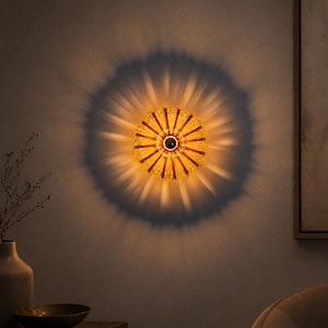 Op de afbeelding: Decoratieve wandlamp met een stervormig ontwerp. De lamp is gemaakt van amberkleurig glas en straalt een warme, uitnodigende gloed uit. Het licht werpt een radiale schaduw op de muur, waardoor een dramatisch effect ontstaat. De armatuur is op een neutraal gekleurde muur gemonteerd.