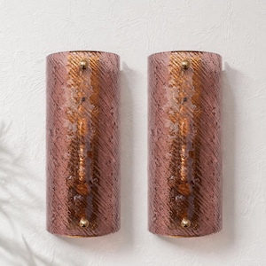 May include: A pair of cylindrical wall sconces in reddish-pink textured glass. Each sconce features a vertical, golden-brown stripe and small brass accents at the top and bottom. They are mounted on a white wall, ideal for interior lighting.