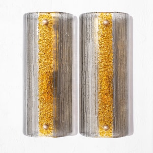 Op de afbeelding: Twee cilindrische wandkandelaars met een textuur, verticaal gestreept ontwerp. Elke kandelaar heeft een centrale band van goudkleurig, getextureerd glas en is geaccentueerd met kleine, ronde, goudkleurige studs. De wandkandelaars zijn op een witte muur gemonteerd.