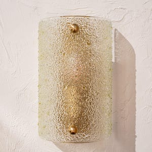 Peut inclure: Applique murale cylindrique avec abat-jour en verre dépoli texturé. L'applique a un cadre en métal doré et deux accents décoratifs ronds dorés. Le luminaire est monté sur un mur blanc texturé.