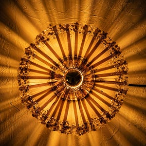 May include: A decorative, circular chandelier with a warm, amber glow. The light fixture features a central, dark-colored disc surrounded by radiating wooden slats. The outer edge is adorned with small, sparkling embellishments, casting a patterned light on the textured wall.