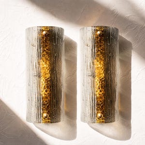 Può includere: Due applique da parete cilindriche con vetro strutturato e dettagli dorati. Ogni applique ha un centro verticale dorato ed è montata su una parete bianca strutturata. Le applique sono illuminate dalla luce solare, creando ombre.
