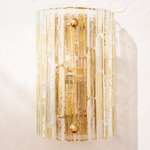 Op de afbeelding: Cilindrische wandlamp met een glazen kap met textuur in goud- en heldere tinten. De lamp heeft een verticaal, baksteenachtig patroon en is bevestigd met twee messingkleurige knoppen. Een decoratieve lamp.