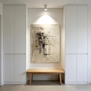May include: An abstract painting with beige, brown, and black brushstrokes hangs above a wooden bench. The artwork is centered on a white wall, flanked by two white storage cabinets. The room is well-lit with a ceiling spotlight.