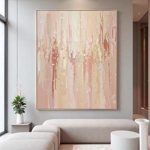 May include: Large abstract painting in a gold frame. The artwork features vertical brushstrokes in shades of peach, cream, and light brown, with scattered white speckles. The painting hangs above a light-colored sofa in a modern living room setting.