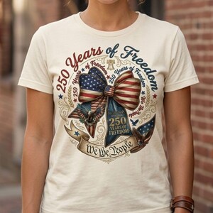 May include: Cream-colored t-shirt featuring a patriotic design. The graphic includes a bow with the American flag, an eagle, and the text "250 Years of Freedom" and "We the People".