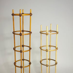 Pode incluir: Dois treliças cilíndricas para plantas feitas de hastes verticais finas conectadas por três anéis circulares horizontais. A treliça mais alta tem um tom de madeira castanho-médio quente, enquanto a mais curta tem uma cor de madeira bege clara.