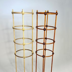 Può includere: Due supporti cilindrici per piante, uno con finitura color crema chiaro e uno in tonalità legno marrone medio. Ogni struttura presenta tre aste verticali collegate da quattro anelli circolari orizzontali, con dettagli a nodo.