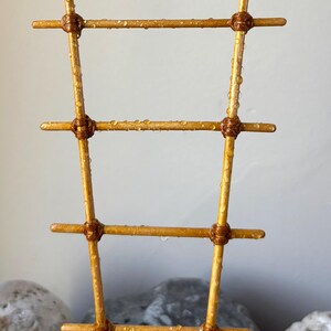 May include: A golden-colored plant trellis featuring four horizontal rungs attached to two vertical poles with dark brown cord wrappings. The structure is covered in small, clear water droplets, giving the surface a wet, glistening appearance.