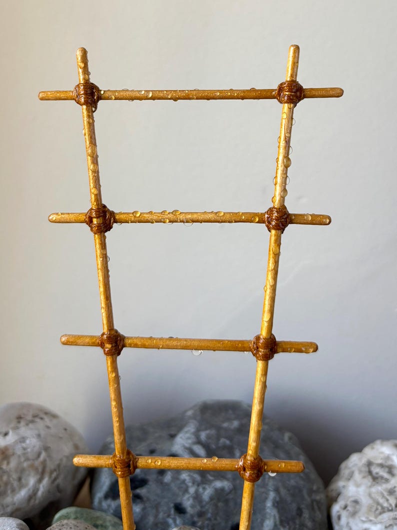 May include: A golden-colored plant trellis featuring four horizontal rungs attached to two vertical poles with dark brown cord wrappings. The structure is covered in small, clear water droplets, giving the surface a wet, glistening appearance.