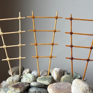 以下が含まれることがあります： 茶色の濃淡の木製トレリスプランターサポートが3つ、滑らかなグレーとベージュの川石の上に配置されています。トレリスは、細い垂直および水平の木片で構成され、紐で接合されています。