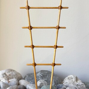 May include: A light brown wooden plant trellis featuring a ladder-like design with four horizontal rungs secured to two vertical supports by thin, wrapped cord detailing at each intersection point, creating a tapered, narrow structure.