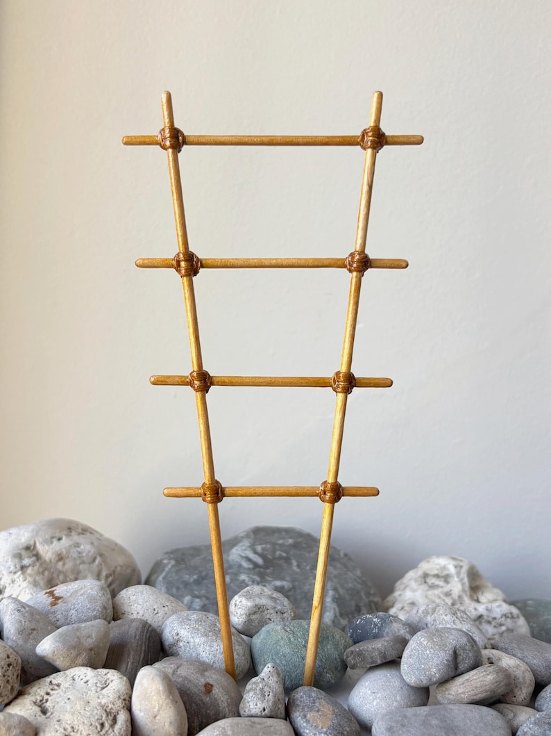 May include: A light brown wooden plant trellis featuring a ladder-like design with four horizontal rungs secured to two vertical supports by thin, wrapped cord detailing at each intersection point, creating a tapered, narrow structure.