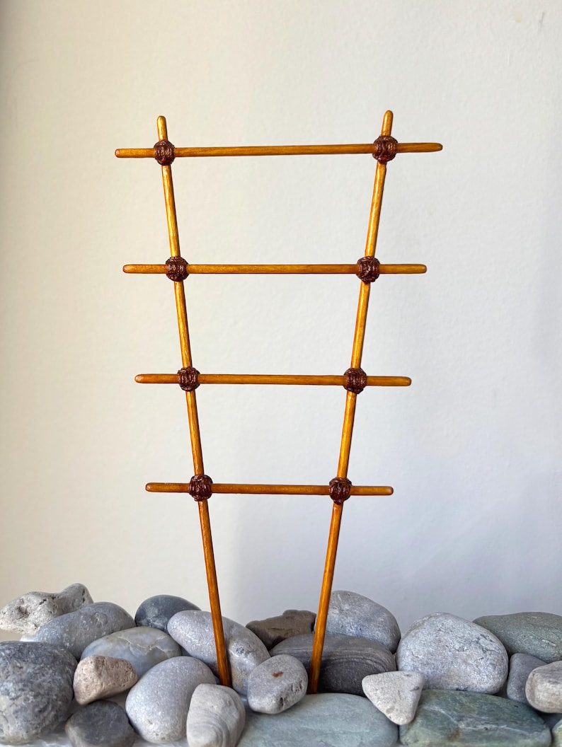 May include: A wooden plant trellis featuring two vertical rods and four horizontal rungs, connected by dark brown knotted cord accents. The structure is light brown with a smooth finish, designed with a tapered shape that is wider at the top.