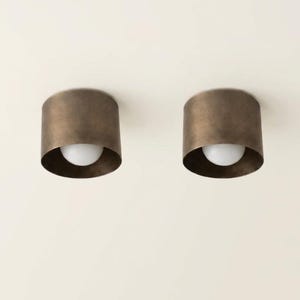 Puede incluir: Dos luces de techo cilíndricas de color bronce con una bombilla blanca visible en la parte inferior. Las luces están montadas en un techo blanco. Las luminarias tienen un diseño simple y moderno.