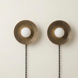 May include: Two wall sconces with a brass finish and a white globe light in the centre. Each sconce has a circular design and a black cord. The sconces are mounted on a white wall.