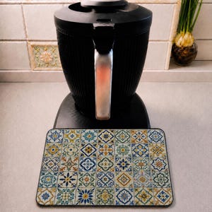 Può includere: Un elettrodomestico da cucina nero con una texture a coste verticali e una maniglia argentata, posizionato dietro un tappetino rettangolare con un motivo ripetuto di piastrelle floreali e geometriche blu, gialle e verdi a griglia.