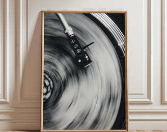 May include: Black and white photograph of a record player in motion. The turntable spins, creating a blurred effect. The tonearm and cartridge are in focus, set against a wooden frame.