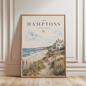 Puede incluir: Una lámina enmarcada que muestra un paisaje costero con una casa de playa en un acantilado, dunas de arena y olas del mar. El texto dice THE HAMPTONS NEW YORK con un pequeño icono de concha centrado bajo el texto en tonos beige claro.