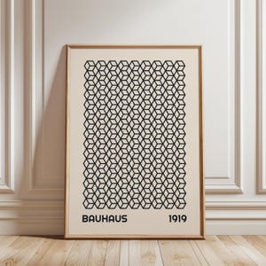 May include: A framed Bauhaus art print. The artwork features a repeating geometric pattern of black cubes on a cream background. The word "BAUHAUS" and the year "1919" are printed in black at the bottom of the print. The frame is a light brown color.