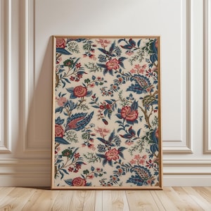 Puede incluir: Una impresión floral enmarcada con un fondo crema, que presenta un patrón repetido de flores y follaje rojo, azul y verde. La impresión está en un marco delgado de color dorado, apoyada contra una pared blanca y un suelo de madera.