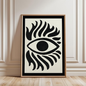 May include: Framed art print featuring a bold, abstract eye design. The artwork showcases a black eye with a circular pupil, surrounded by stylized flame-like shapes, set against a cream background. The print is framed in a thin, natural wood frame.