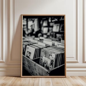 May include: Black and white photograph of a framed print featuring a collection of vinyl records in a wooden crate. The records are in their sleeves, with some album art visible. The image is in a room with white paneling and a wooden floor.