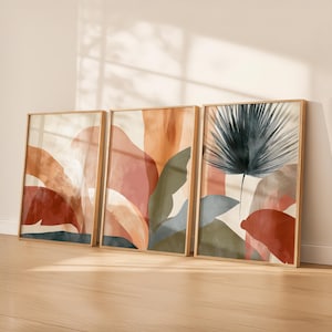 May include: Three framed abstract art prints. The artwork features organic shapes and botanical elements in shades of rust, peach, green, and blue. The frames are a light wood color, and the prints are displayed against a white wall.