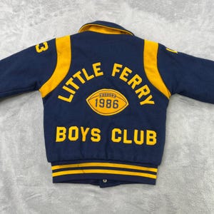 May include: A navy blue and yellow Little Ferry Boys Club varsity jacket. The jacket features yellow accents on the sleeves and collar, with the words "LITTLE FERRY" and "BOYS CLUB" in yellow lettering. A yellow football graphic with the year "1986" is also visible.