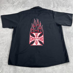 May include: A black short-sleeve button-down shirt featuring a large graphic on the back. The graphic displays a white iron cross with red outlines and red flames rising from the top, containing the text West Coast Choppers in red letters.