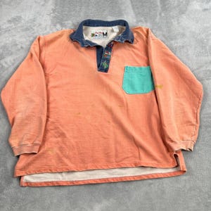 May include: A long-sleeve peach-colored pullover shirt with a dark blue denim-style collar and placket. It features a single teal chest pocket, a white inner collar label with the text "Ping Pong" and "M", and a small decorative flag patch.