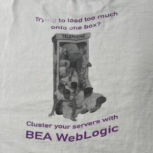May include: A white t-shirt featuring a purple graphic of a telephone booth overflowing with people. The text reads: Trying to load too much onto one box? Cluster your servers with BEA WebLogic. The design is printed in a dark purple ink on white fabric.