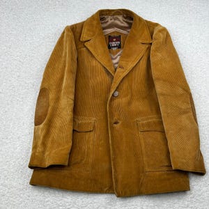 May include: A corduroy blazer in a warm, golden-brown hue. Features include a notched lapel, two front pockets, and suede elbow patches. The jacket has a single-button closure and a label that reads "EUROPE CRAFT".