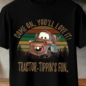 May include: A black short-sleeve t-shirt featuring a graphic of a rusty brown tow truck character. The design includes a sunset background with horizontal stripes and the text "COME ON, YOU'LL LOVE IT!" and "TRACTOR-TIPPIN'S FUN." in a tan font.