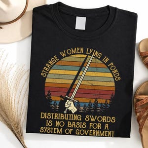 May include: A black t-shirt featuring a graphic of a hand holding a sword against a retro-style sunset with pine trees. The text reads: STRANGE WOMEN LYING IN PONDS DISTRIBUTING SWORDS IS NO BASIS FOR A SYSTEM OF GOVERNMENT in a gold-colored font.