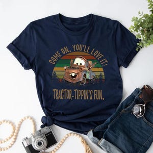 May include: A navy blue short-sleeve t-shirt featuring a graphic of a brown tow truck character. The design includes a sunset background with horizontal stripes and the text "COME ON, YOU'LL LOVE IT!" and "TRACTOR-TIPPIN'S FUN." in a tan font.