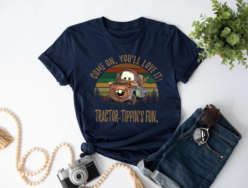 May include: A navy blue short-sleeve t-shirt featuring a graphic of a brown tow truck character. The design includes a sunset background with horizontal stripes and the text "COME ON, YOU'LL LOVE IT!" and "TRACTOR-TIPPIN'S FUN." in a tan font.