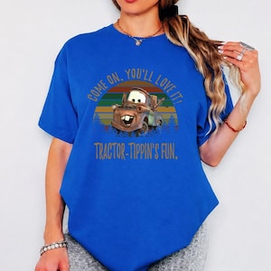 May include: A bright blue short-sleeve t-shirt featuring a graphic of the character Mater from the movie Cars. The design includes a sunset background and the text: COME ON, YOU'LL LOVE IT! TRACTOR-TIPPIN'S FUN. The shirt has a crew neckline.