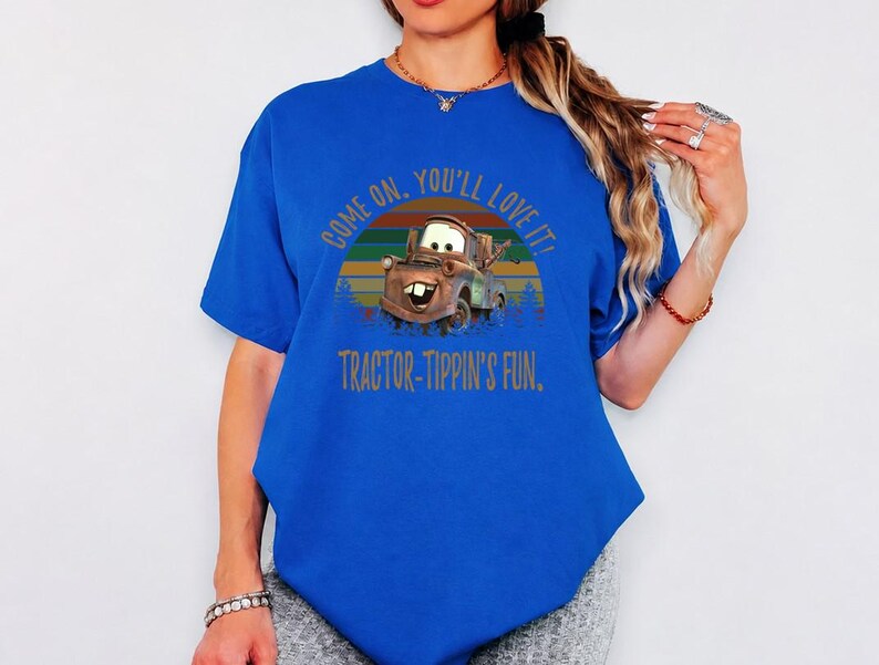 May include: A bright blue short-sleeve t-shirt featuring a graphic of the character Mater from the movie Cars. The design includes a sunset background and the text: COME ON, YOU'LL LOVE IT! TRACTOR-TIPPIN'S FUN. The shirt has a crew neckline.