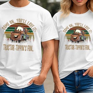 May include: A white short-sleeved t-shirt featuring a graphic of a brown tow truck character. The design includes a sunset background with green and orange stripes, pine trees, and the text: COME ON, YOU'LL LOVE IT! TRACTOR-TIPPIN'S FUN.