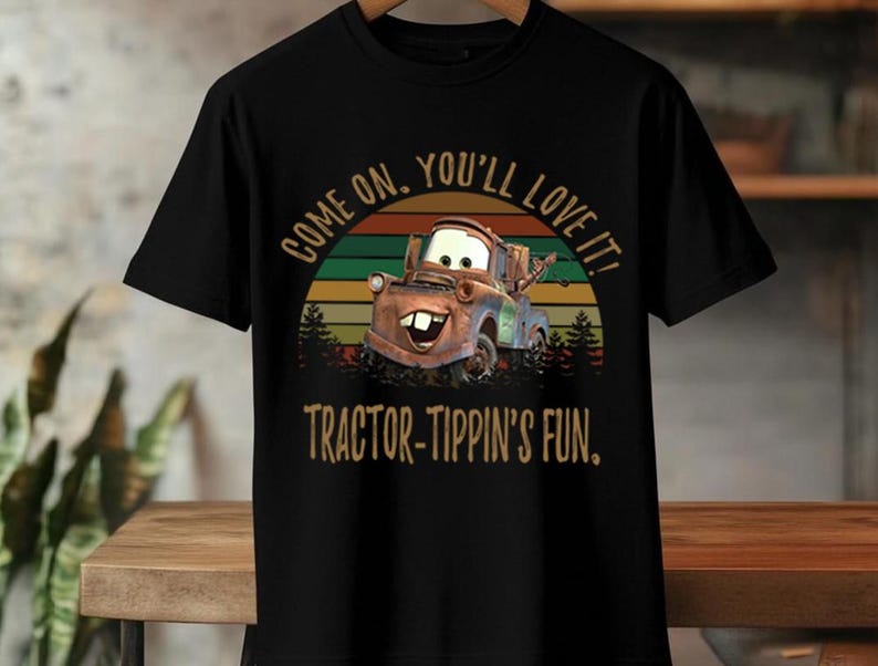 May include: A black short-sleeve t-shirt featuring a graphic of a rusty brown tow truck character. The design includes a sunset background with horizontal stripes and the text "COME ON, YOU'LL LOVE IT!" and "TRACTOR-TIPPIN'S FUN." in a tan font.