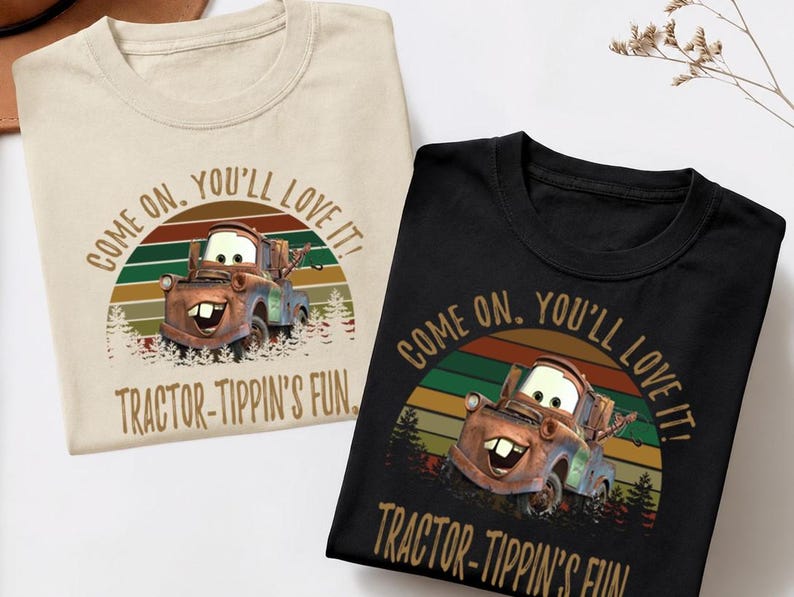 May include: A beige t-shirt and a black t-shirt, each featuring a graphic of a rusty tow truck character. The design includes a sunset background with horizontal stripes, pine trees, and the text "COME ON, YOU'LL LOVE IT! TRACTOR-TIPPIN'S FUN."