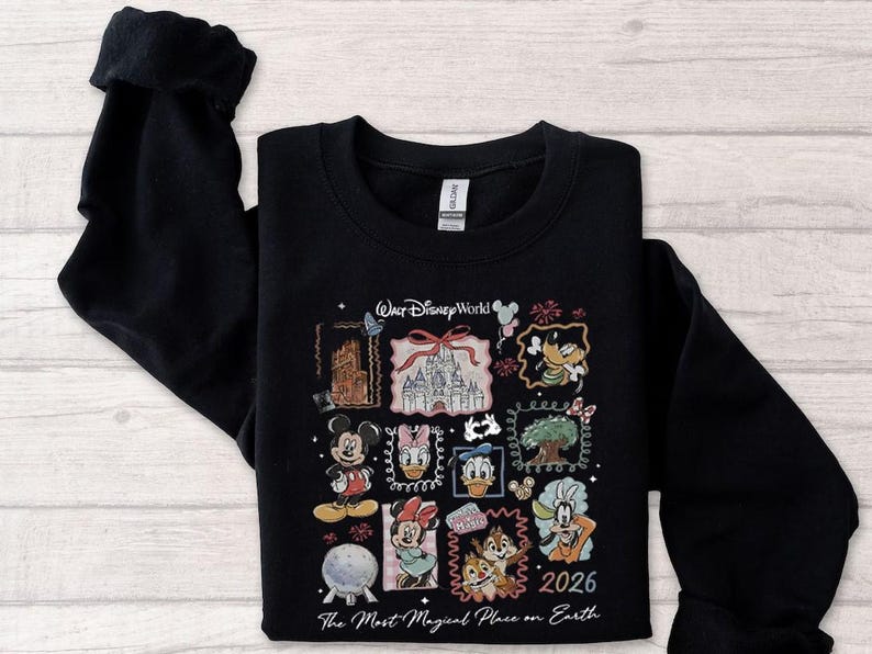 May include: Black sweatshirt featuring a colorful graphic of Disney characters and landmarks. The design includes Mickey Mouse, Donald Duck, and Cinderella's castle, with the text "Walt Disney World" and "2026". The phrase "The Most Magical Place on Earth" is also included.