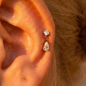 Op de afbeelding: Een goudkleurige oorbelset. De bovenste stud heeft een ronde, heldere edelsteen. Een druppelvormige edelsteen hangt eronder. De oorbel wordt close-up getoond, waarbij de details worden benadrukt.